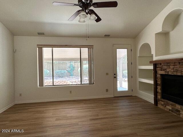 unfurnished living room featuring a stone fireplace, built in shelves, hardwood / wood-style floors, and ceiling fan