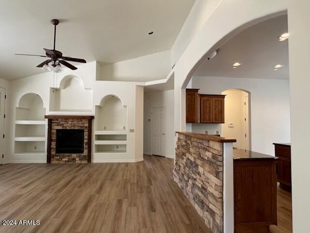 unfurnished living room with ceiling fan, lofted ceiling, a stone fireplace, hardwood / wood-style flooring, and built in shelves