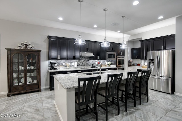 kitchen with light stone countertops, hanging light fixtures, an island with sink, a breakfast bar, and appliances with stainless steel finishes