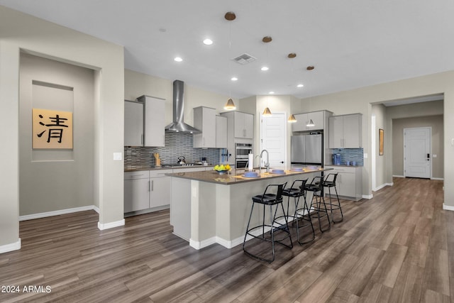 kitchen with pendant lighting, visible vents, appliances with stainless steel finishes, a kitchen island with sink, and wall chimney range hood