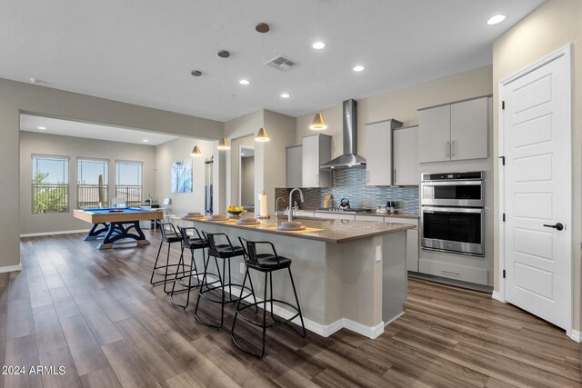 kitchen featuring stainless steel appliances, an island with sink, pool table, wall chimney range hood, and dark wood-type flooring