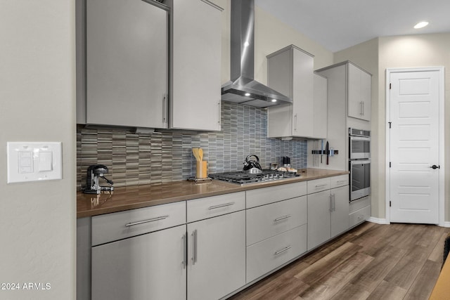 kitchen with wall chimney range hood, appliances with stainless steel finishes, tasteful backsplash, dark countertops, and dark wood finished floors