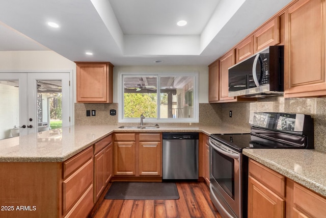 kitchen featuring a sink, a tray ceiling, dark wood-style floors, a peninsula, and appliances with stainless steel finishes