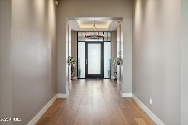 hallway featuring a tray ceiling, a chandelier, baseboards, and light wood finished floors