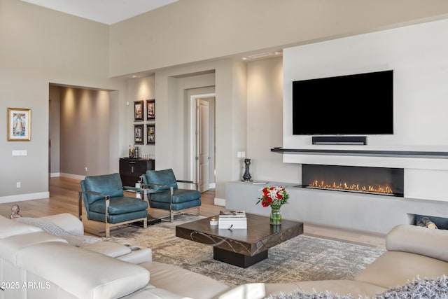 living area featuring wood finished floors, baseboards, a lit fireplace, and a high ceiling