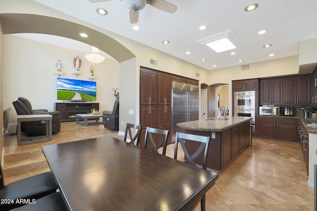 kitchen with dark brown cabinets, stainless steel appliances, a kitchen island with sink, sink, and stone counters