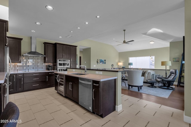 kitchen featuring appliances with stainless steel finishes, wall chimney exhaust hood, sink, ceiling fan, and a center island with sink