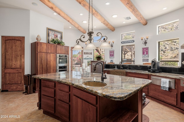 kitchen with a center island with sink, decorative light fixtures, light stone countertops, sink, and beamed ceiling