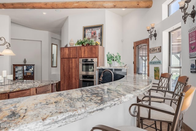 kitchen featuring a high ceiling, light stone counters, beamed ceiling, double oven, and decorative light fixtures