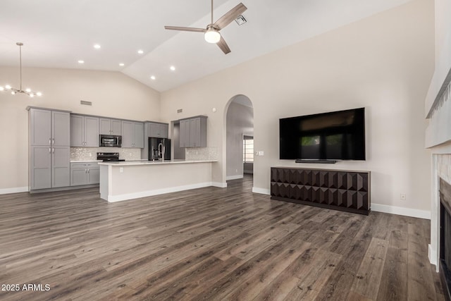 unfurnished living room with high vaulted ceiling, dark wood-type flooring, ceiling fan with notable chandelier, arched walkways, and a fireplace