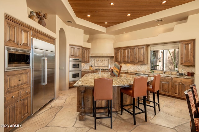 kitchen featuring wood ceiling, light stone countertops, built in appliances, and an island with sink