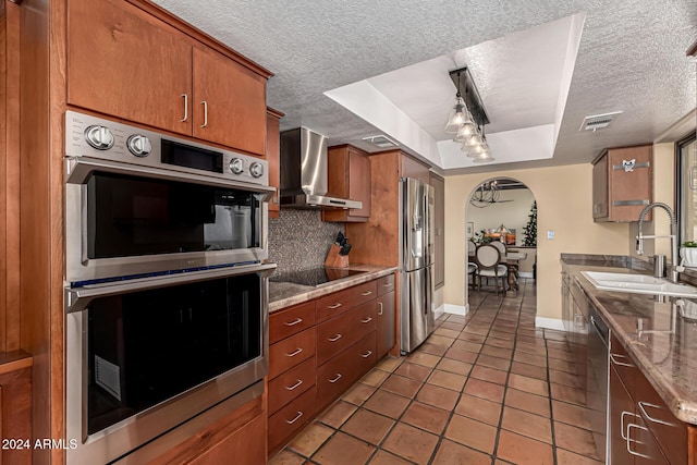 kitchen with sink, wall chimney range hood, a textured ceiling, a tray ceiling, and appliances with stainless steel finishes