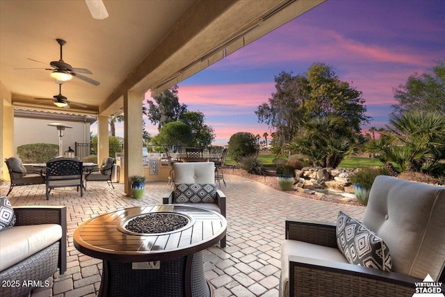 patio terrace at dusk with an outdoor kitchen, ceiling fan, and an outdoor living space with a fire pit