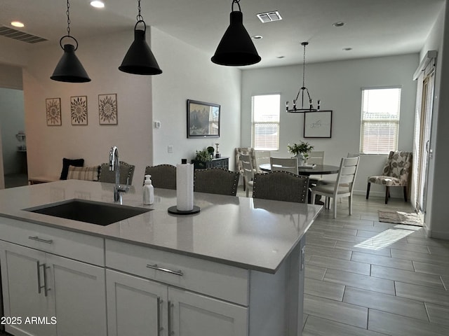 kitchen featuring a center island with sink, sink, hanging light fixtures, light stone counters, and white cabinetry