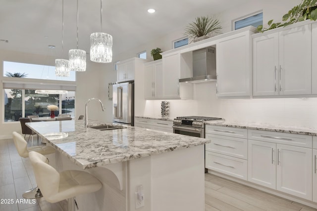 kitchen featuring high end appliances, hanging light fixtures, white cabinetry, a sink, and wall chimney range hood