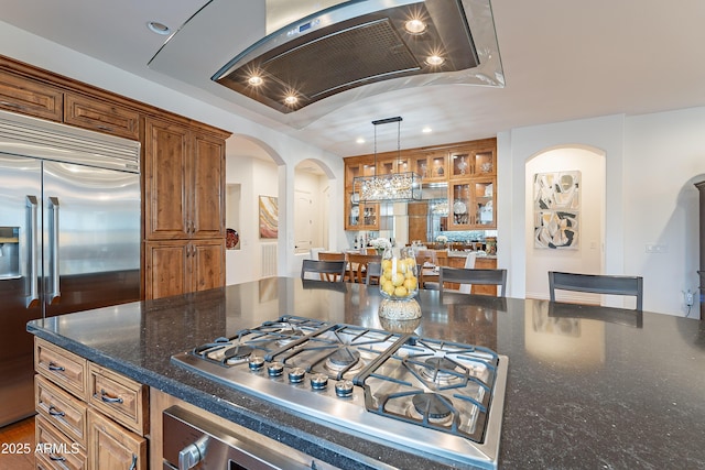 kitchen with arched walkways, brown cabinetry, glass insert cabinets, appliances with stainless steel finishes, and pendant lighting