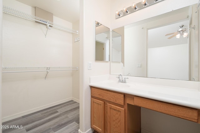 bathroom with a walk in closet, wood finished floors, baseboards, ceiling fan, and vanity