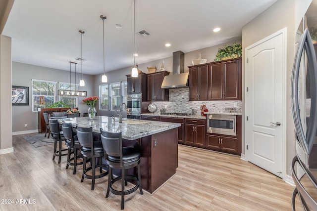 kitchen with light stone countertops, wall chimney exhaust hood, stainless steel appliances, a breakfast bar area, and a center island with sink