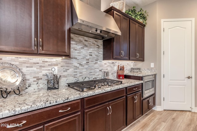 kitchen featuring dark brown cabinetry, light stone countertops, wall chimney exhaust hood, stainless steel appliances, and light hardwood / wood-style flooring