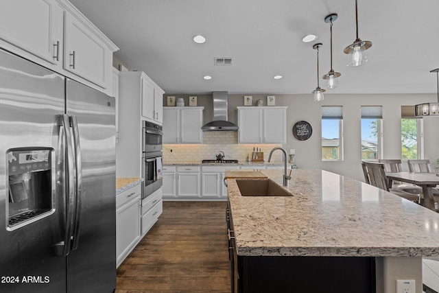 kitchen with wall chimney range hood, appliances with stainless steel finishes, pendant lighting, sink, and a kitchen island with sink