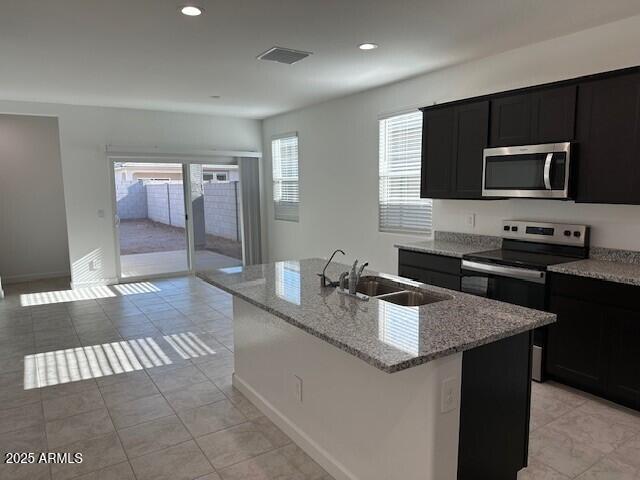 38055 W Excussare Way, Maricopa, AZ 85138 - kitchen with appliances with stainless steel finishes, dark cabinetry, a sink, light stone counters, and a kitchen island with sink