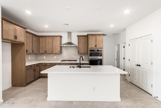 kitchen featuring wall chimney range hood, a sink, appliances with stainless steel finishes, tasteful backsplash, and an island with sink