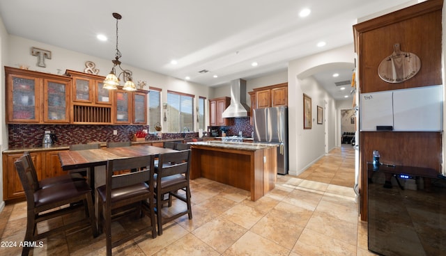 kitchen featuring tasteful backsplash, decorative light fixtures, a center island, stainless steel fridge, and wall chimney range hood