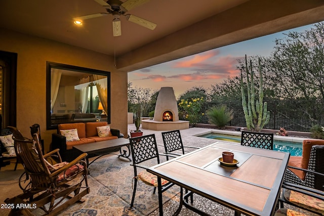 patio terrace at dusk with an outdoor living space with a fireplace, ceiling fan, and a fenced in pool