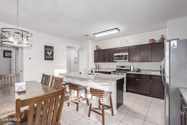 kitchen with appliances with stainless steel finishes, light stone counters, dark brown cabinets, a center island with sink, and hanging light fixtures