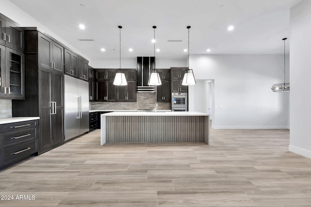 kitchen featuring stainless steel appliances, range hood, backsplash, decorative light fixtures, and a kitchen island with sink