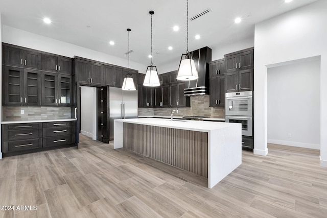 kitchen with pendant lighting, sink, a kitchen island with sink, stainless steel appliances, and wall chimney range hood