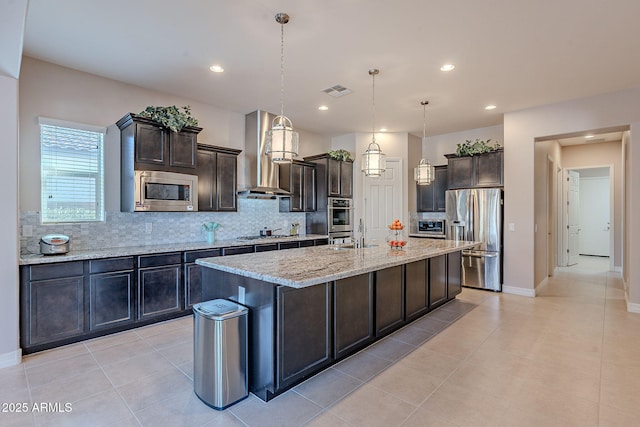 kitchen featuring appliances with stainless steel finishes, hanging light fixtures, dark brown cabinets, an island with sink, and wall chimney exhaust hood