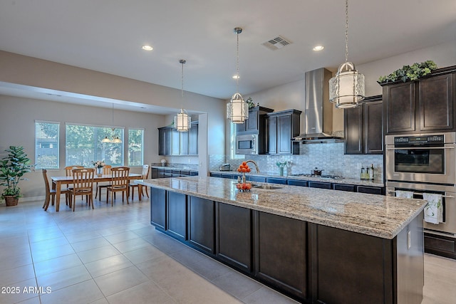 kitchen featuring wall chimney exhaust hood, sink, decorative light fixtures, appliances with stainless steel finishes, and a kitchen island with sink