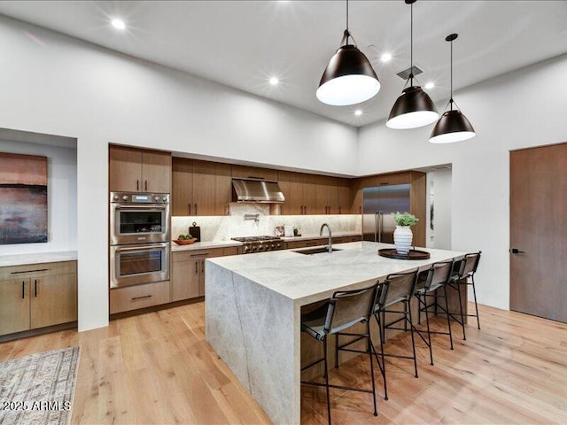 kitchen with sink, stainless steel appliances, light hardwood / wood-style floors, decorative light fixtures, and a large island with sink