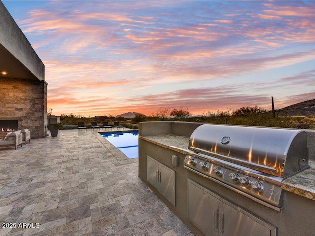 patio terrace at dusk featuring a grill, area for grilling, and an outdoor stone fireplace