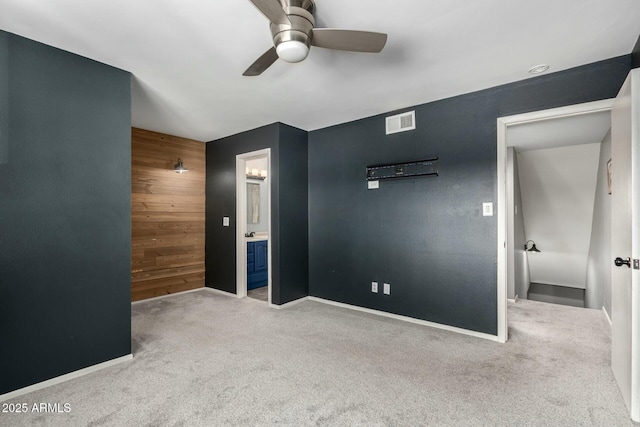 unfurnished bedroom with light carpet, ensuite bath, wooden walls, and ceiling fan