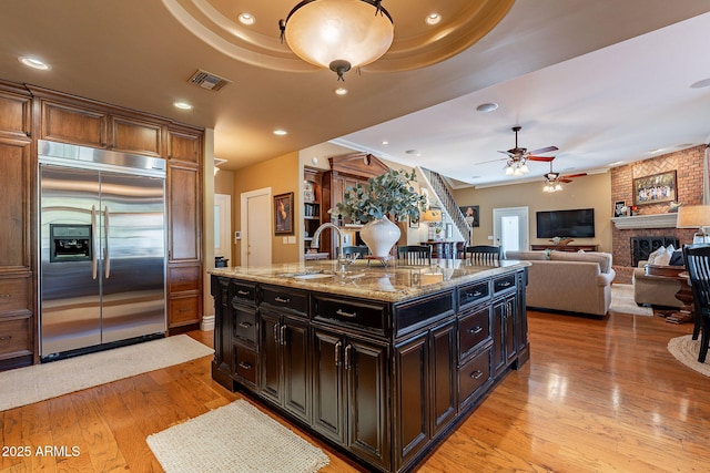 kitchen with sink, a brick fireplace, a center island with sink, stainless steel built in fridge, and light stone countertops