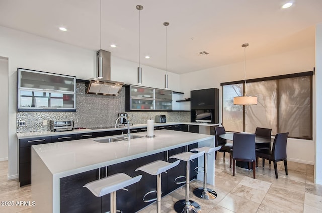 kitchen featuring backsplash, a kitchen island with sink, pendant lighting, and wall chimney range hood