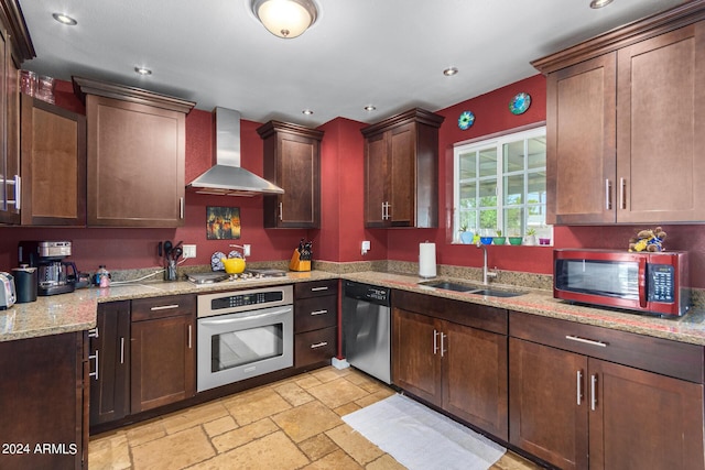 kitchen featuring stone tile floors, appliances with stainless steel finishes, a sink, wall chimney range hood, and light stone countertops