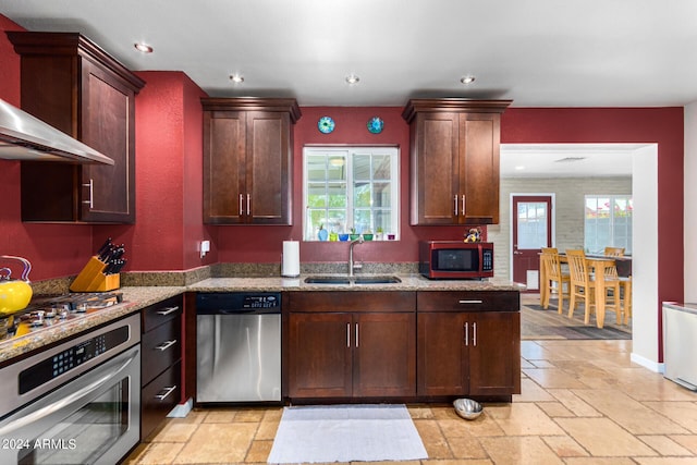 kitchen featuring light stone counters, a sink, appliances with stainless steel finishes, wall chimney exhaust hood, and stone tile flooring