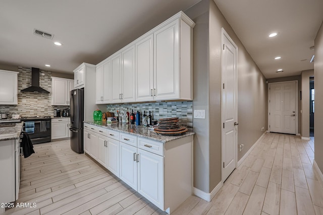 kitchen with black refrigerator, white cabinetry, stove, light stone counters, and wall chimney exhaust hood