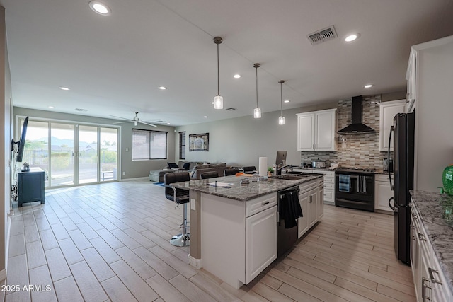 kitchen featuring hanging light fixtures, black appliances, an island with sink, white cabinets, and wall chimney exhaust hood