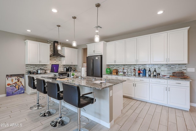 kitchen with white cabinets, stainless steel fridge, hanging light fixtures, light stone countertops, and wall chimney range hood