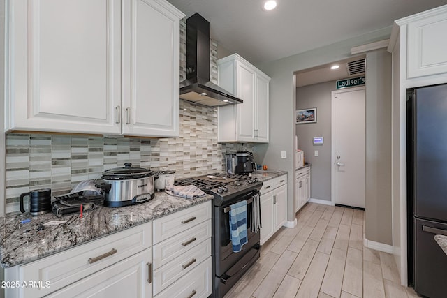 kitchen featuring stainless steel refrigerator, white cabinetry, black range with gas stovetop, light stone counters, and wall chimney range hood