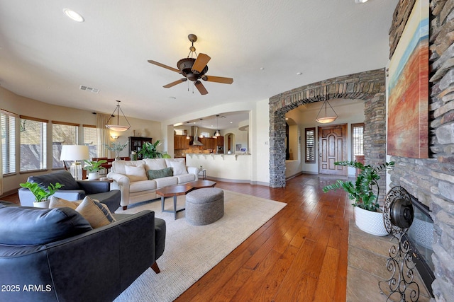 living room featuring visible vents, arched walkways, baseboards, wood-type flooring, and a stone fireplace