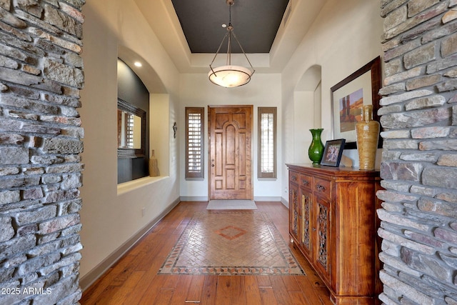 foyer entrance featuring wood-type flooring, arched walkways, a raised ceiling, and baseboards