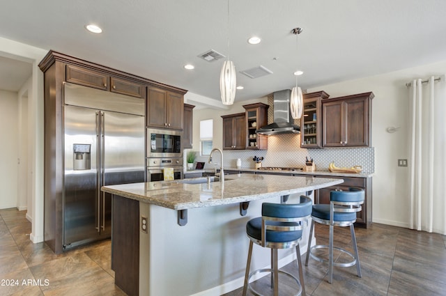 kitchen with sink, wall chimney exhaust hood, built in appliances, pendant lighting, and a center island with sink