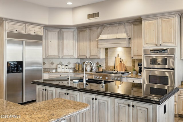 kitchen with premium range hood, stainless steel appliances, cream cabinets, and dark stone countertops