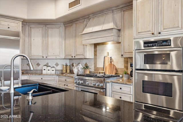 kitchen with sink, appliances with stainless steel finishes, backsplash, custom exhaust hood, and dark stone counters