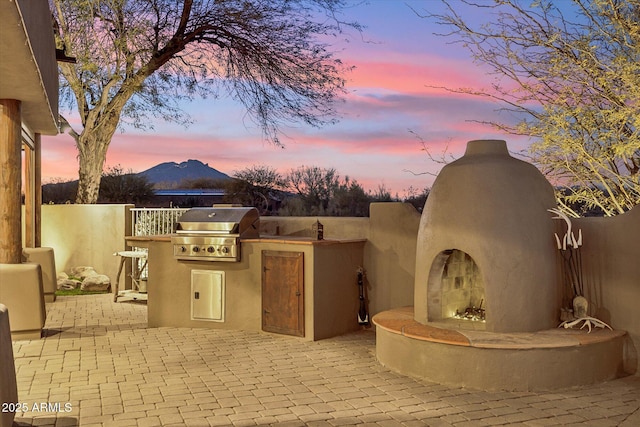 patio terrace at dusk featuring exterior fireplace, grilling area, a mountain view, and exterior kitchen